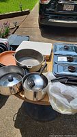Overview of all cookware displayed on a table outdoors including saucepans, pot, heating pans and cutting boards.