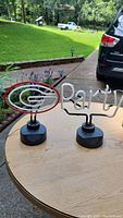 Two neon lights, one is the University of Georgia logo in red and white neon tubing and the other spells 'Party' in white neon tubing, both mounted on black bases, placed on a wooden table outdoors.