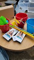 Red and blue plastic buckets with attached handles, bright green shovels and leaf-shaped sand molds, and yellow and orange plastic water guns on a round wooden table.