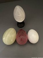 Four glass eggs on a dark surface, with the frosted clear egg engraved with a partridge displayed upright on a black stand, next to the greenish-white swirled egg, solid white egg, and deep reddish-pink egg.