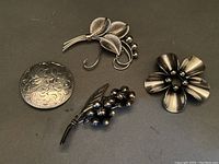 Four sterling silver brooches showing different styles and designs including floral and berry motifs on a dark background.