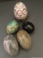 Four stone eggs in turquoise, black, beige, and gray/pink tones and one ornate porcelain egg box with floral pattern and gilded hinge and clasp on a grey surface.