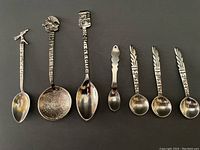 Seven sterling silver Mexican souvenir spoons laid out for display on a dark background showing their unique handle designs and varied bowl shapes.