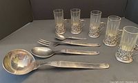 Photo showing ladle, salad servers, teaspoon, and six tea glasses in silver holders arranged on a surface.
