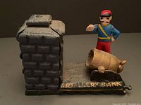 Side view showing the artillery officer in blue jacket and red pants with the cannon, on the black base labeled 'ARTILLERY BANK'. The coin slot and lever mechanism on the cannon are visible.