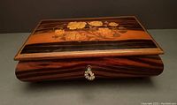 Front angle view of closed rectangular wooden musical jewelry box showing floral inlaid lid and keyhole.