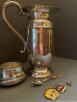 Silver plate pitcher with hinged lid and ornate handle, souvenir spoon, three pins laid out on dark surface