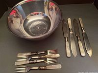 Silver plated large bowl with clear plastic liner, accompanied by four fish forks and four fish knives with mother of pearl handles on black background.