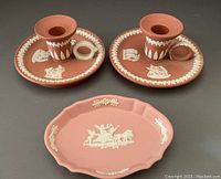 Front view photo showing two terracotta Wedgwood Jasperware chamber-stick candle holders and one pink oval Wedgwood Jasperware pin dish all with white relief decoration.