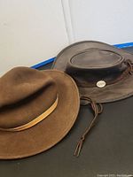 Two pre-owned hats on black surface, one brown wool felt fedora, one brown leather Western-style hat with chin strap