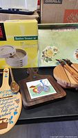 Photo showing the 10-inch Joyce Chen Bamboo Steamer box, wooden spatula with painted text, wooden cutting boards with tile insets, round wooden trivet/coaster, and decorative floral box.
