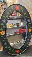Full view of the oval mirror showing hand painted colorful flowers on the black frame with some scratches visible.