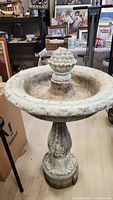 Front angled view of concrete bird bath showing decorative finial and weathered surface with dirt and wear.