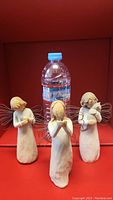 Three Willow Tree angel figurines displayed on a red shelf next to a water bottle for scale, showing full body front view.