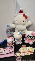 Photo showing large white Hello Kitty plush in Santa hat, ceramic clock with blue bow, small plush toys, pink Nintendo 3DS case, and Hello Kitty playing cards spread out.
