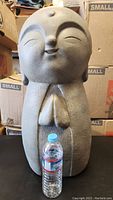 Full view of the Jizo statue with a water bottle for scale, showing the statue's peaceful smiling face and prayer hands.