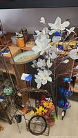 Photo showing miscellaneous patio decor items including brass bird bath, solar flower spinner, metal pot flower holder, white artificial floral spray, grapevine wreath with flowers, two metal garden trellises, and solar greenish glass globe.