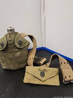 Full view of vintage US Army canteen in green canvas holder, beige canvas belt beside it, and small matching green pouch with snap-button.