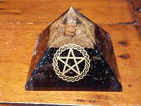 Close-up photo of the orgonite pyramid showing the black base with gold flecks, clear top with copper coil, and gold pentagram symbol on front face.