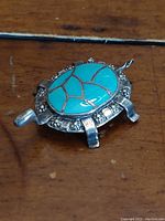 Front view of turquoise and sterling silver turtle-shaped pendant/brooch showing the turquoise stone inlaid with silver lines and surrounding decorative silver detailing.