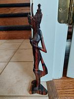 Side view of the slender wooden Thai dancer figurine showing the intricate headdress and crossed arm pose on a small base.