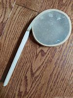 Photo of the front side of the vintage handcrafted rattle showing the round hide skin rattling area and white wooden handle on wood floor background.
