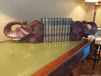 Pair of dark reddish-brown wooden elephant head bookends holding a row of vintage-style books on a green and brown table.