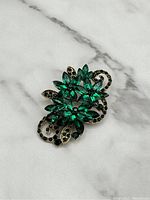 Emerald green and black stone brooch on gold tone metal with floral and swirl design, shown on marble background