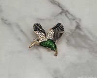 Photo of a hummingbird brooch with emerald green enamel body, black enamel and clear rhinestones on wings, gold-tone accents, on a white marble background.