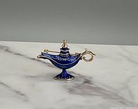 Close-up photo of a gold-tone brooch shaped like a genie lamp with blue enamel and decorative detailing on a white marble surface.