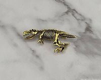 Front view of the gold tone dinosaur skeleton brooch pin, showcasing detailed bone and skull features on a marble-like background.