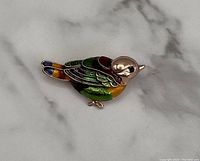 Photo of gold tone bird brooch with multicolor enamel, showing detailed side view on a marble surface.