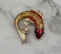 Close-up photo of a gold tone koi fish brooch showing red and orange enamel detailing on the body, textured gold tone scales, flowing gold metal fins, and green stone eyes.