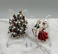 Two gold tone pins displayed on clear stands, including a leaf design with rhinestones and a dancing couple with multicolor enamel and faux pearl detail.