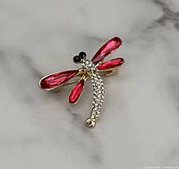 Close-up photo of the gold tone dragonfly brooch with red translucent wings and clear rhinestones on body, black rhinestone eyes, on a marble surface.