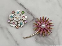 Photo of two gold tone flower brooches on marble surface showing multicolored rhinestones and faux pearl detail.