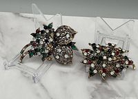 Two floral design brooches with multicolor rhinestones displayed on clear holders on a white surface