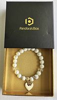 White and gold beaded bracelet with heart-shaped charm displayed inside a black box with gold Pandoras Box logo.