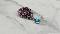 Side angle of brooch showing pink, red, purple gemstones and blue dangling stone on silver-tone metal setting