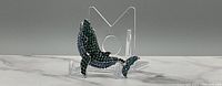 Side angle of silver-tone whale brooch showing green and blue rhinestone decoration on a marble surface with a clear acrylic stand in the background.