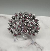 Front view of silver tone peacock brooch with pink and black stones on white marble surface