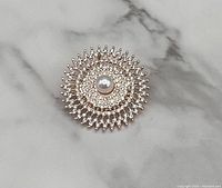 Top view of brooch showing round design, decorated with rose gold tone metal and clear rhinestones around a central faux pearl.