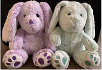 Two vintage plush bunnies, one purple and one pale green, sitting side by side showing front view with paw pads visible.