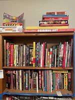 Three-tier bookshelf showing two upper shelves of mixed comics and art books and top shelf featuring two Simpsons-themed games