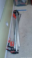 Six Bessey 39-inch bar clamps laid on the floor, showing metal bars and red handles.