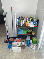Full view of cleaning supplies including bottles, towels stacked on shelves, broom, dustpan, and other supplies on floor.