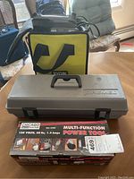 Photo showing Ryobi yellow and black carrying bag, gray hard Dremel case, and Chicago Electric boxed multi-function power tool on table.