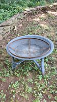 Full view of round metal fire pit on three legs, situated outdoors on grass and stones.