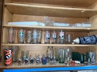 Full shelf view showing multiple glasses, steins, shot glasses, Coca Cola pitcher, flask and bottle openers arranged on wooden shelves.