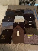 25 men’s T-shirts in various colors and sizes laid out flat on a bed.